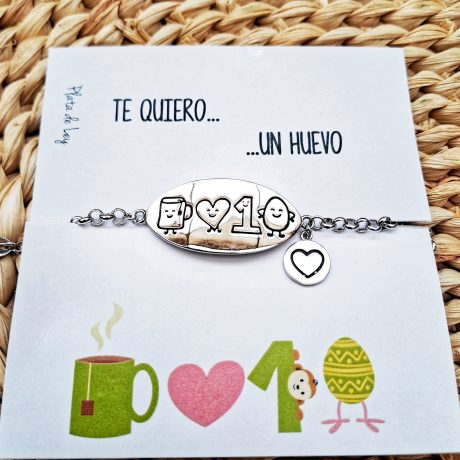 pulsera de plata con una cadena y en el centro una chapa ovalada con esta grabado a laser el dibujo de una taza, un corazon, un numero uno y un huevo, con una pequeña chapita redonda a un lado donde esta graabada un corazon